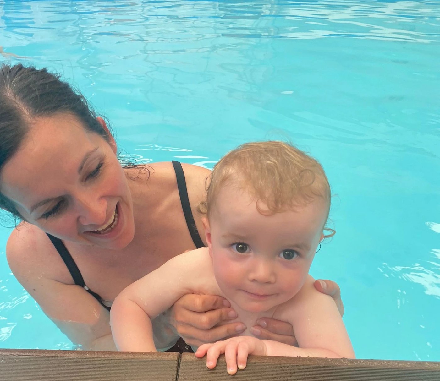 A smiling mum supporting her baby at the poolside as the baby holds the edge and looks at the camera
